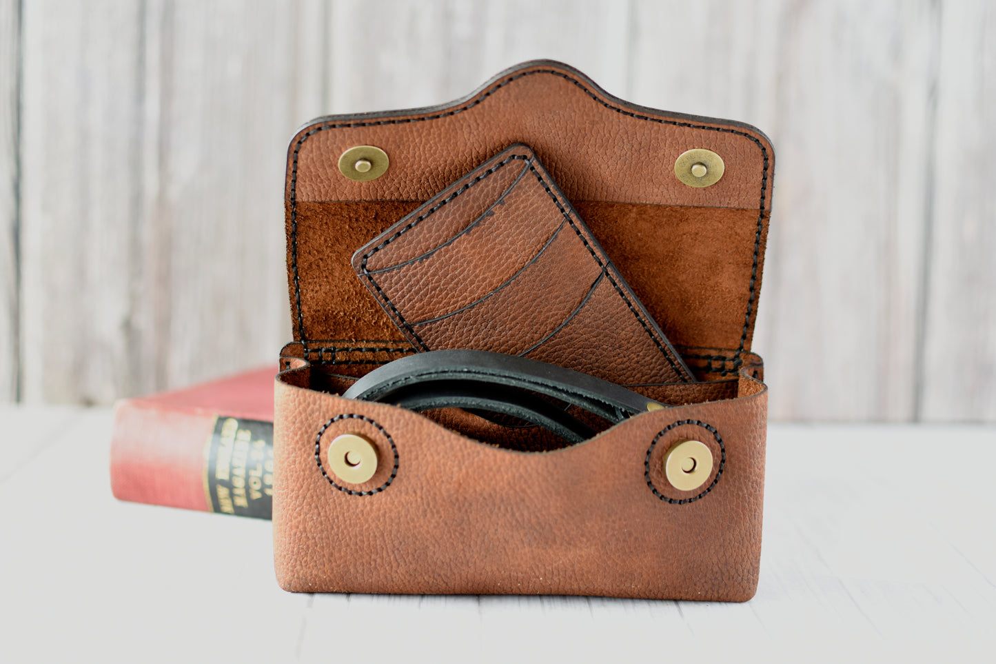 Brown leather cell phone purse on a wooden surface with a book in the background with compact wallet and black strap